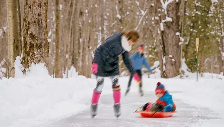 A person pulls a child on a sled as they skate across a country trail surrounded by fluffy white snow banks on either side.