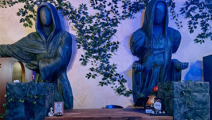 Kunstinstallation im Freien mit zwei vermummten Skulpturenfiguren, die an einem von Laubwerk umgebenen Tisch sitzen.