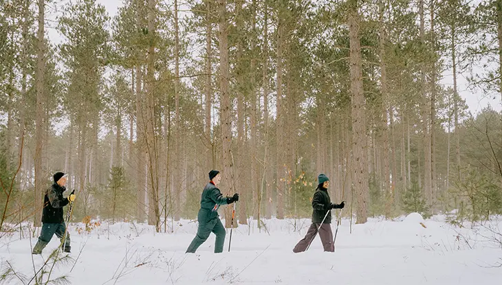 Tres personas practican esquí de fondo en un tranquilo bosque con altos pinos, disfrutando de una apacible excursión invernal.