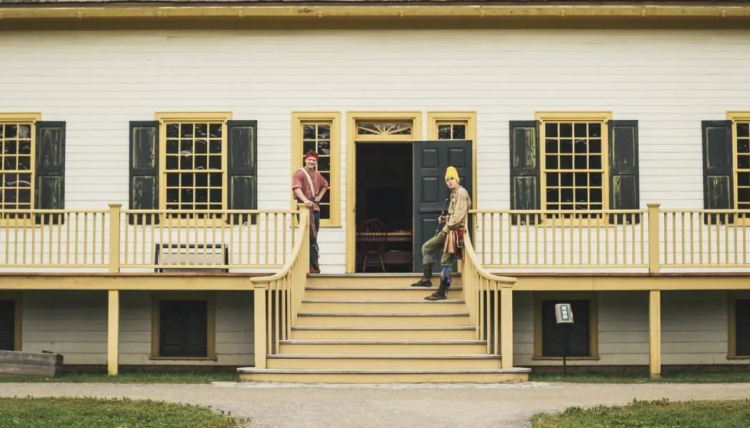Two men dressed in Voyageur fur trade costumes stand at the top of a set of stairs leading to the front door of a building at Fort William Historical Park in Thunder Bay.