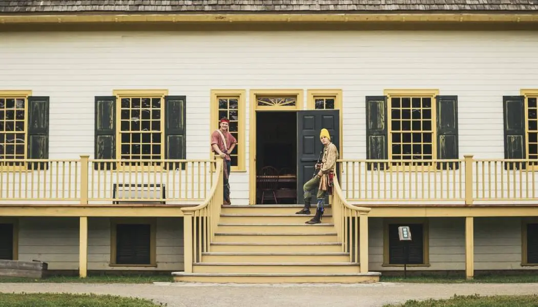 Deux hommes en costume de voyageur se tiennent à l’entrée d’un grand bâtiment en bois au parc historique du Fort William, un musée d’histoire vivante à Thunder Bay.