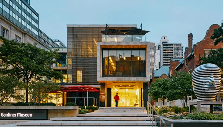 Entrada moderna al museo con fachada de cristal, escalones anchos y gente entrando al anochecer.