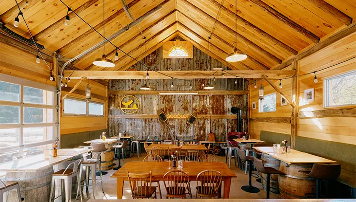 A cozy brewery pub with wood tables, hanging lights and a lit-up sign with "Goodlot" in the background.
