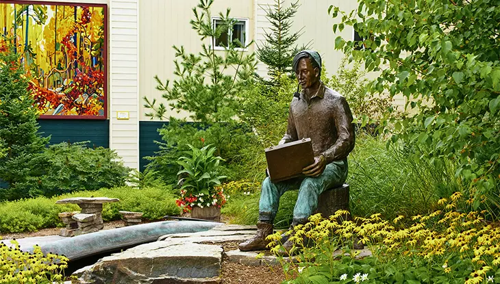 Escultura de bronce de un artista sentado sosteniendo un cuaderno de bocetos en un jardín paisajístico afuera de una galería de arte, con una pintura colorida exhibida en el fondo.