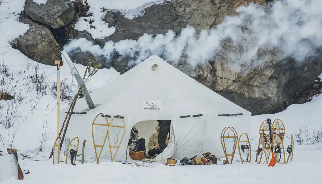A winter camping site with smoke rising from the chimney of a hot tent and several pairs of snowshoes in the snow.