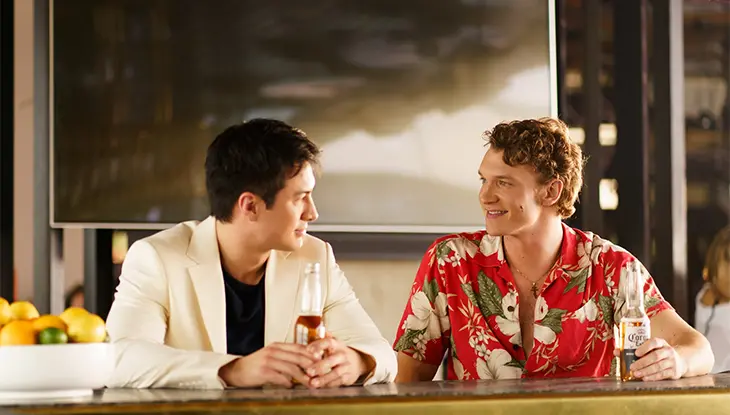 Two men sit at a bar holding beer bottles and chatting, one wearing a white blazer and the other a red floral shirt.