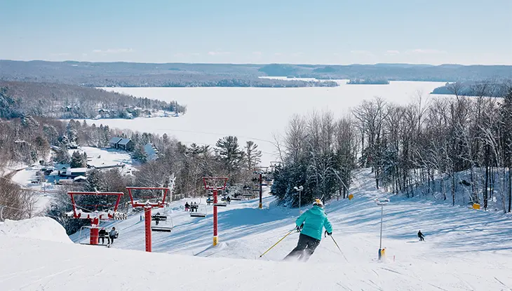 Skieur descendant une pente enneigée qui surplombe un lac gelé, avec un paysage forestier et un remonte-pente au loin.