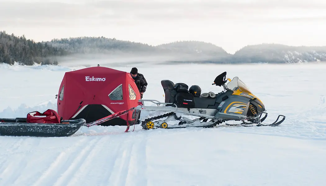 Un hombre instala un refugio para pescar en el hielo de un lago congelado, junto a su moto de nieve y su trineo.