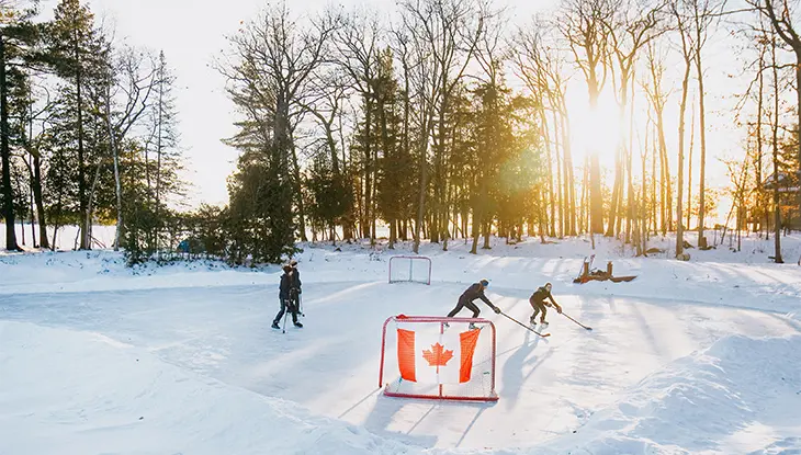 People playing pond hockey on an outdoor ice rink in winter with trees and sunset in the background.