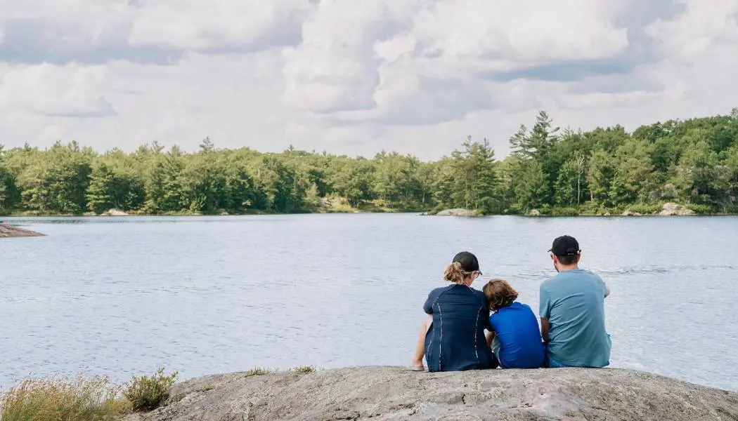 A family of three sits on a rock overlooking a lake surrounded by forest in the Kawarthas.