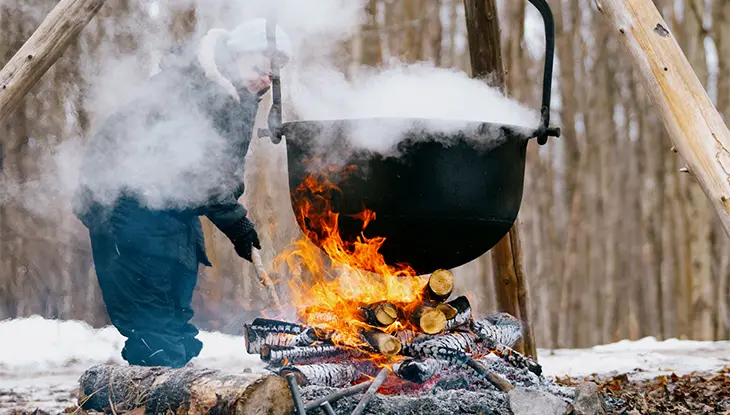 Traditional outdoor maple syrup boiling over an open wood fire, with a large hanging cauldron steaming in a winter forest setting.