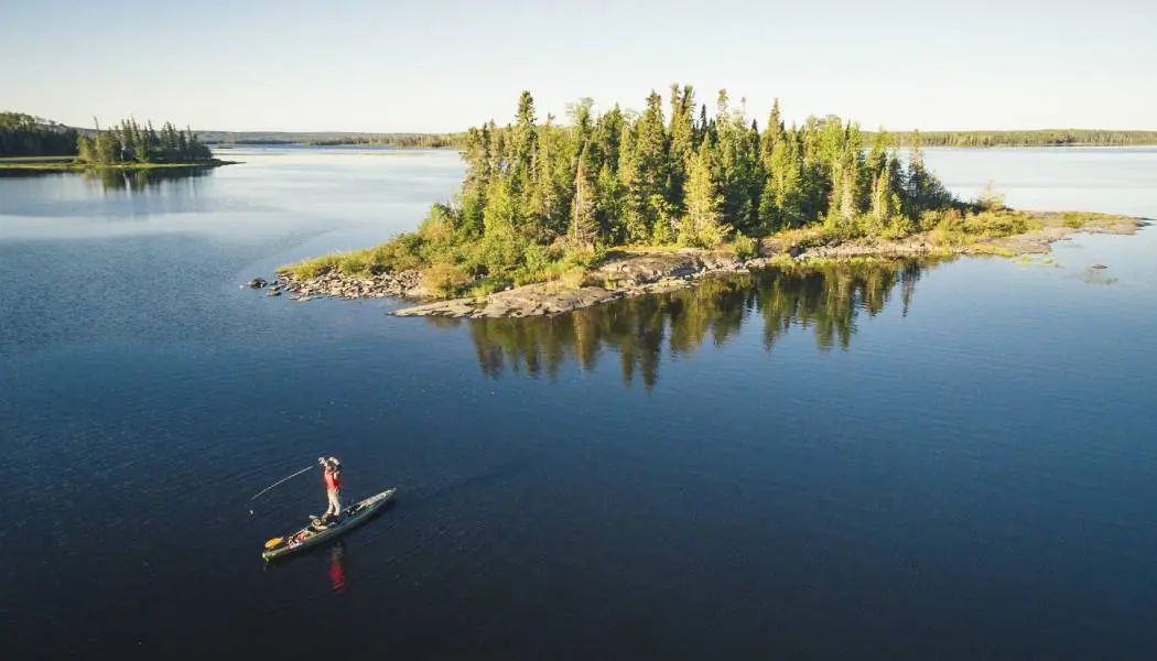 An angler stands up to cast from a fishing kayak on a calm lake near a lush island in Northwest Ontario.