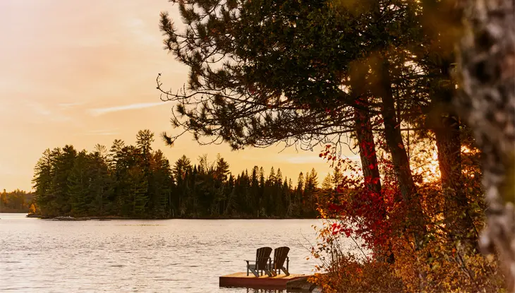 Dos sillas Adirondack vacías en Killarney Lodge, Algonquin Park, en un muelle de madera con vista a un lago tranquilo con el cálido resplandor del sol poniente.