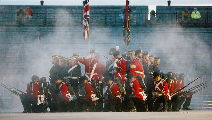 Reconstituteurs historiques en uniforme rouge avec des fusils au milieu de la fumée, exécutant un exercice militaire avec des drapeaux.