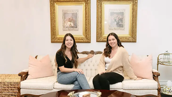 Kristen y Kathryn Groom, copropietarias de The Rose Manor, sentadas en un sofá color crema en una elegante habitación con obras de arte enmarcadas en oro detrás de ellas.