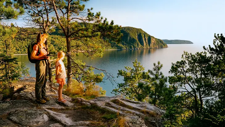 Ein Erwachsener und ein Kind stehen auf einem felsigen Aussichtspunkt im Lake Superior Provincial Park und blicken über die bewaldeten Klippen und den ruhigen See darunter.