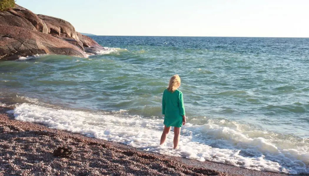 A woman stands in beach surf, where the blue waters of Lake Superior meet a rocky shoreline.
