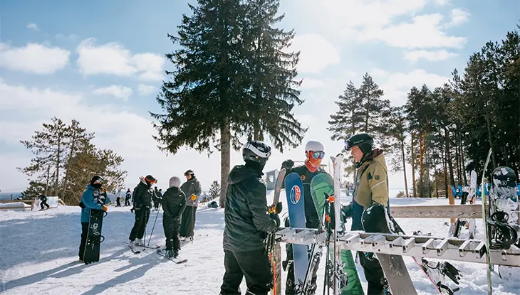 Skieurs et planchistes rassemblés près d’un support à équipement au sommet d’une pente entourée d’arbres, par une journée d’hiver ensoleillée.