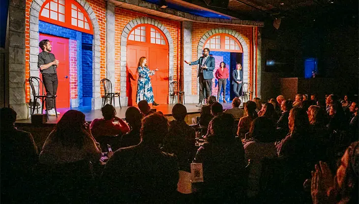 Artistas en el escenario durante un espectáculo de comedia en vivo dentro de un teatro con una audiencia sentada al frente.