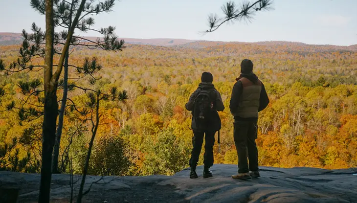 Two hikers on a rocky lookout stop to admire the view of a forest below in full fall colour in Algonquin Park.