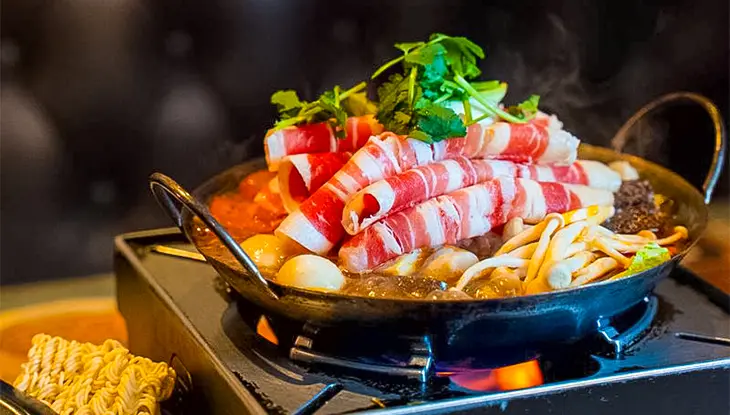 Plat de fondue chinoise avec bœuf tranché, légumes et champignons cuisant sur un brûleur portatif.