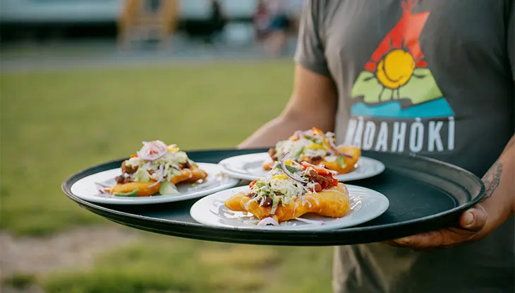 Person holding a tray of plated Indigenous dishes topped with fresh ingredients outdoors.
