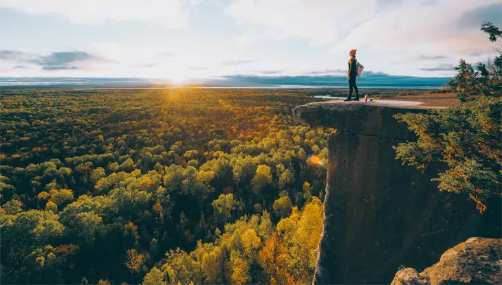 A hiker stands on a rocky lookout over a vast orange and red forest.