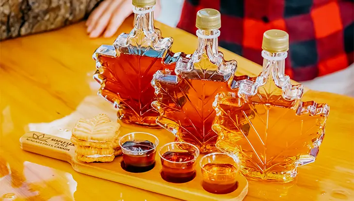 Leaf-shaped bottles of maple syrup arranged on a wooden table with tasting samples and cookies, showcasing different syrup colours and grades.