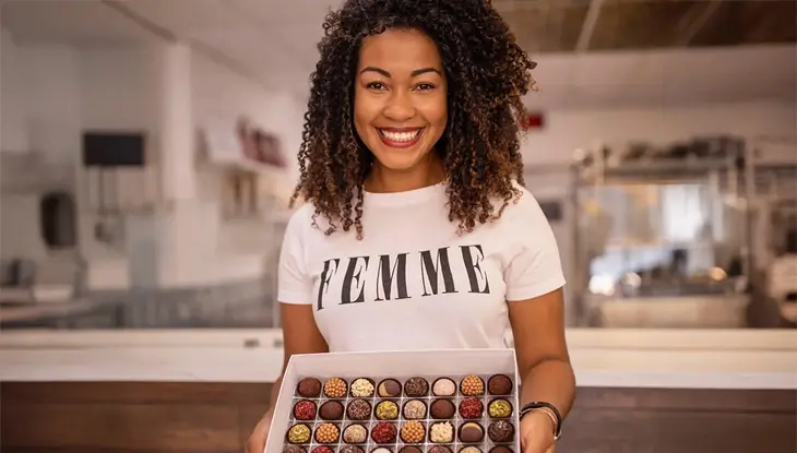 Mariane Oliveira from Brigadeiro wearing a “FEMME” t-shirt holding an open box of assorted chocolates in a bakery setting.