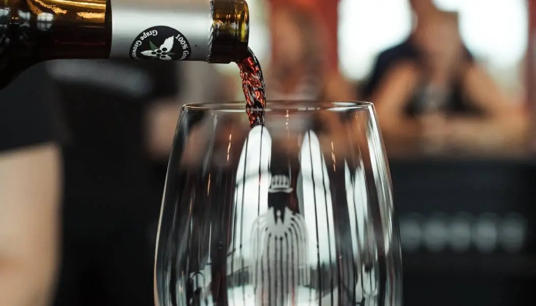 Close-up view of red wine poured into a wine glass with patrons sitting at a tasting bar in the background