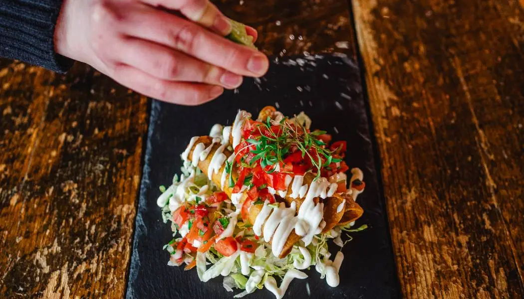 Une personne presse un citron frais sur un taco coloré de taille généreuse, garni de divers ingrédients et de sauce.