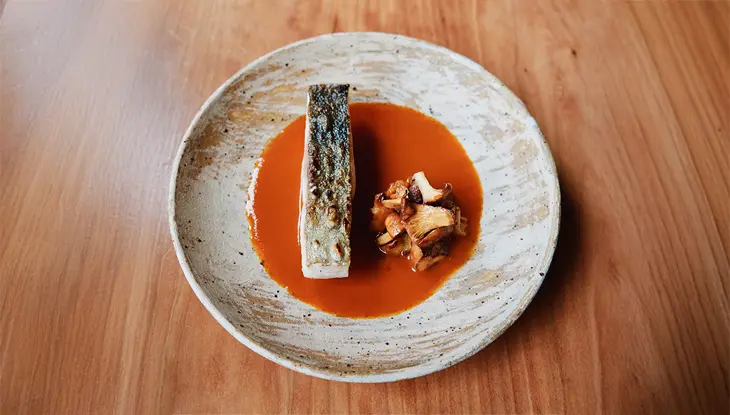 Filet de poisson grillé et croustillant, servi à la verticale dans une sauce rouge liquide, accompagné de champignons sautés, sur un plat en céramique posé sur une table.