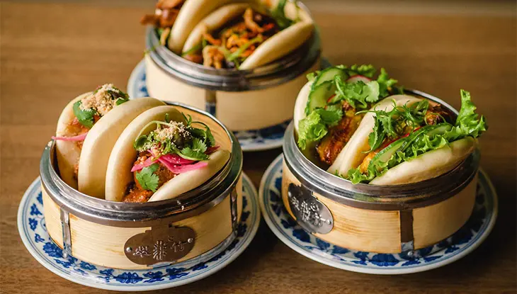 Tres vaporeras de bambú bellamente dispuestas y llenas de bollos bao inflados y sus delicados aderezos de carne y verduras.