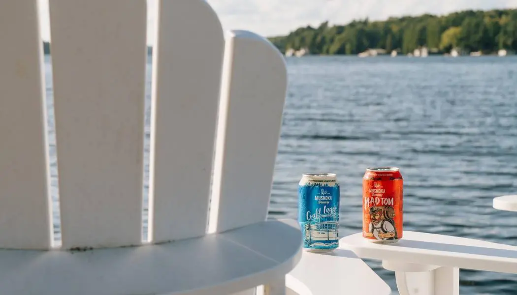 Two craft beers are placed on the armrests of white Muskoka deck chairs overlooking a lake.