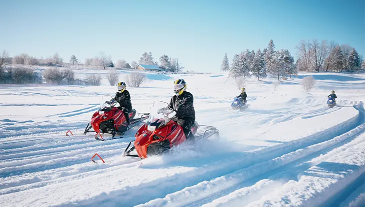 Cuatro personas viajan en motos de nieve por un paisaje nevado iluminado por el sol, creando senderos dinámicos en la nieve.