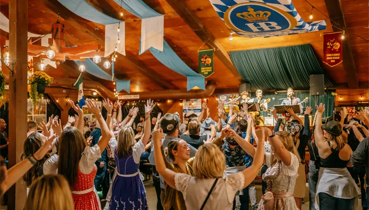 A crowd dressed in Oktoberfest apparel holding their hands up in a lively wooden house while performers sing on stage.