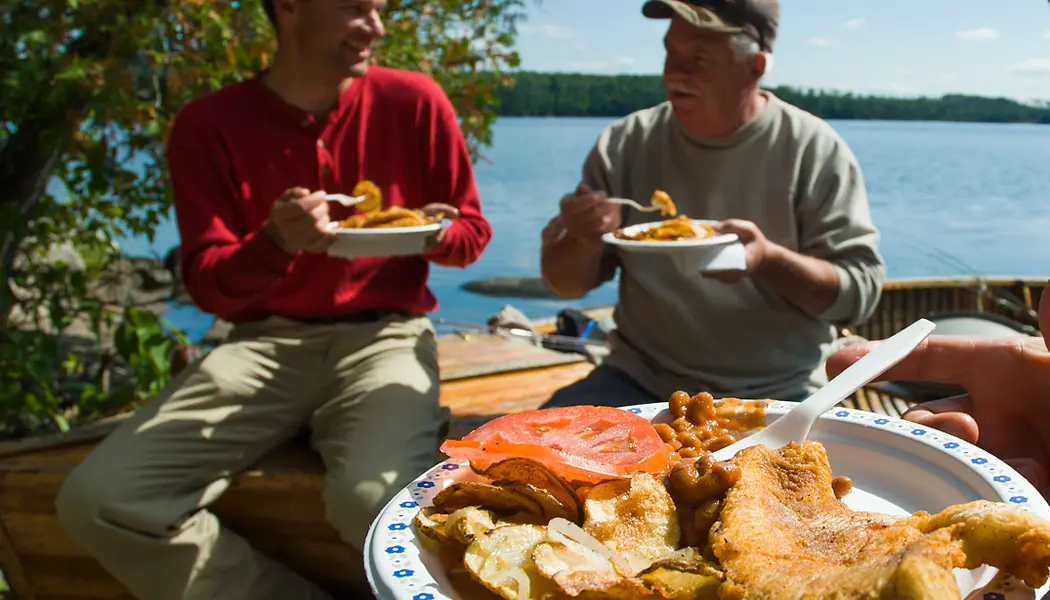 En una excursión de pesca en Ontario, los pescadores disfrutan de un almuerzo a la orilla del mar con pescado fresco cocinado a la brasa, servido con frijoles y verduras.