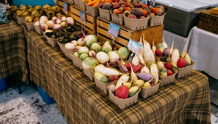 Baskets of assorted root vegetables displayed on a table at an indoor farmers market stall.