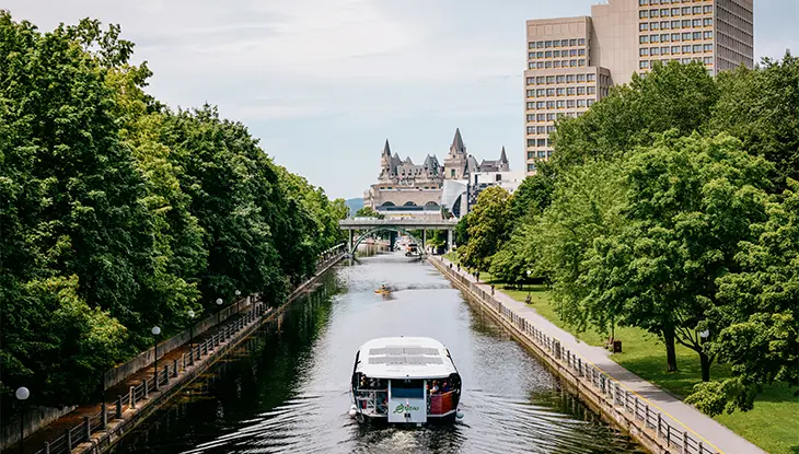 Ein Ausflugsboot fährt auf dem Rideau-Kanal in Ottawa, im Hintergrund sind Château Laurier und Gebäude der Innenstadt zu sehen.
