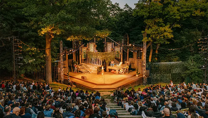 A crowd gathered at an outdoor amphitheatre surrounded by trees, watching a play unfold on a warmly lit stage at dusk.