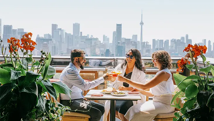 Three people enjoy drinks and snacks on a rooftop looking at the CN and other tall buildings.