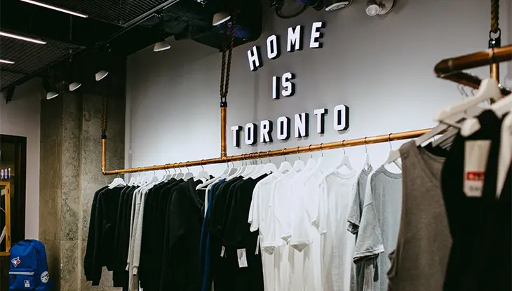 매장 내부에는 검정, 흰색, 회색으로 된 옷걸이가 놓여 있습니다. 뒷벽에는 "Home is Toronto"라는 문구가 적힌 간판이 밝게 빛납니다.