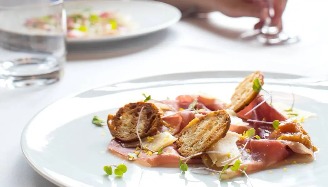 Two colourful, artistically plated summer dishes sit on a white tablecloth with a person’s hand holding the base of a wine glass.