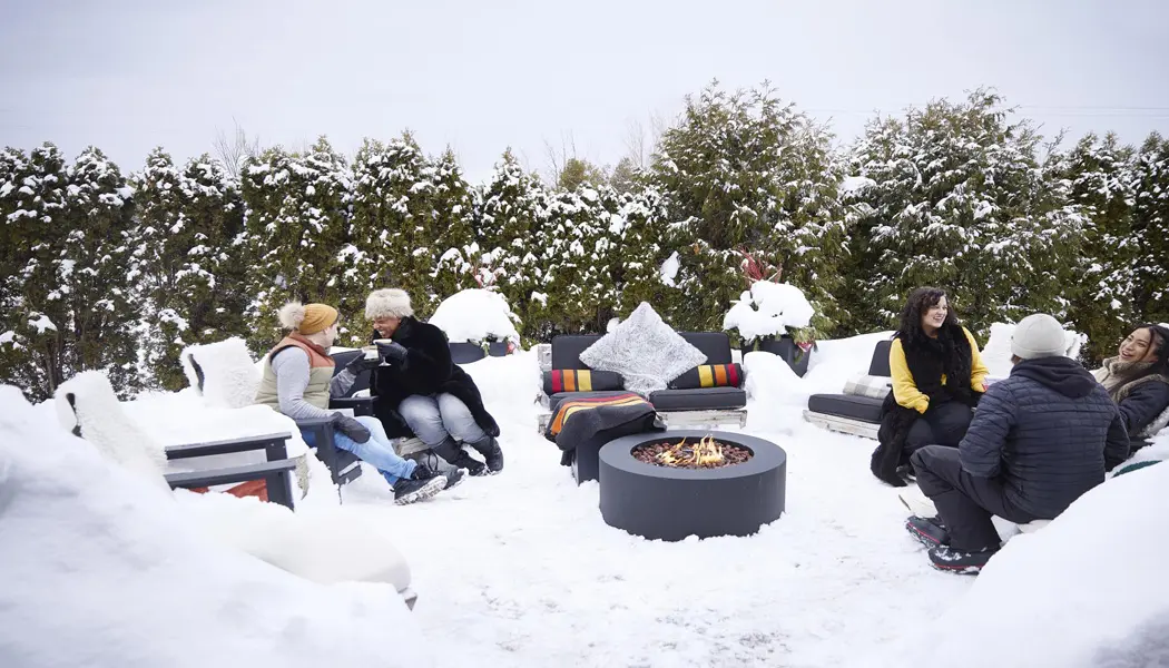 Guests sit outdoors around a fire pit on cozy cushioned chairs with blankets, surrounded by fresh snow and forest.