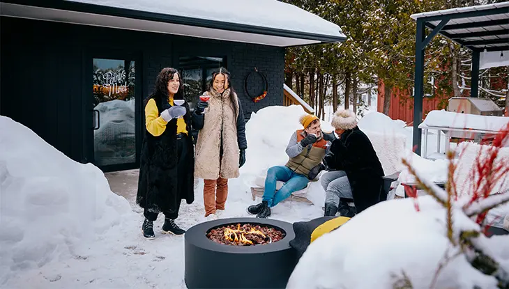 Two stylishly-dressed women hold colourful cocktails outside, while a couple sits with drinks, getting warm by the fire.