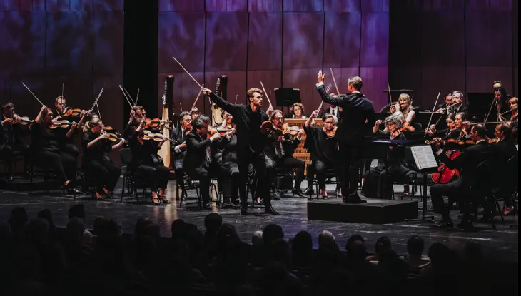 Orchestre complet se produisant dans une salle de concert éclairée par des lumières colorées sur scène.
