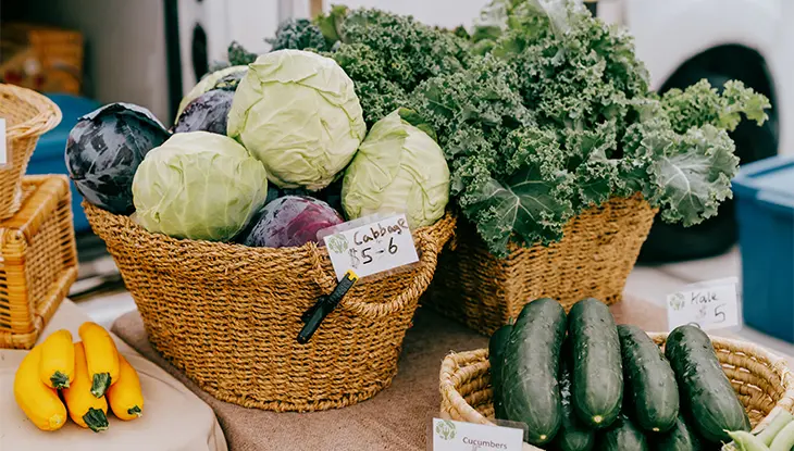Cestas repletas de repollo, col rizada y pepinos frescos expuestas en un puesto de un mercado de agricultores.