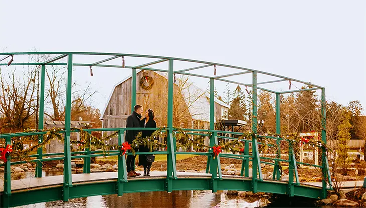 Una pareja tiene un momento romántico en un puente verde cubierto de decoraciones navideñas y un antiguo granero de madera al fondo.