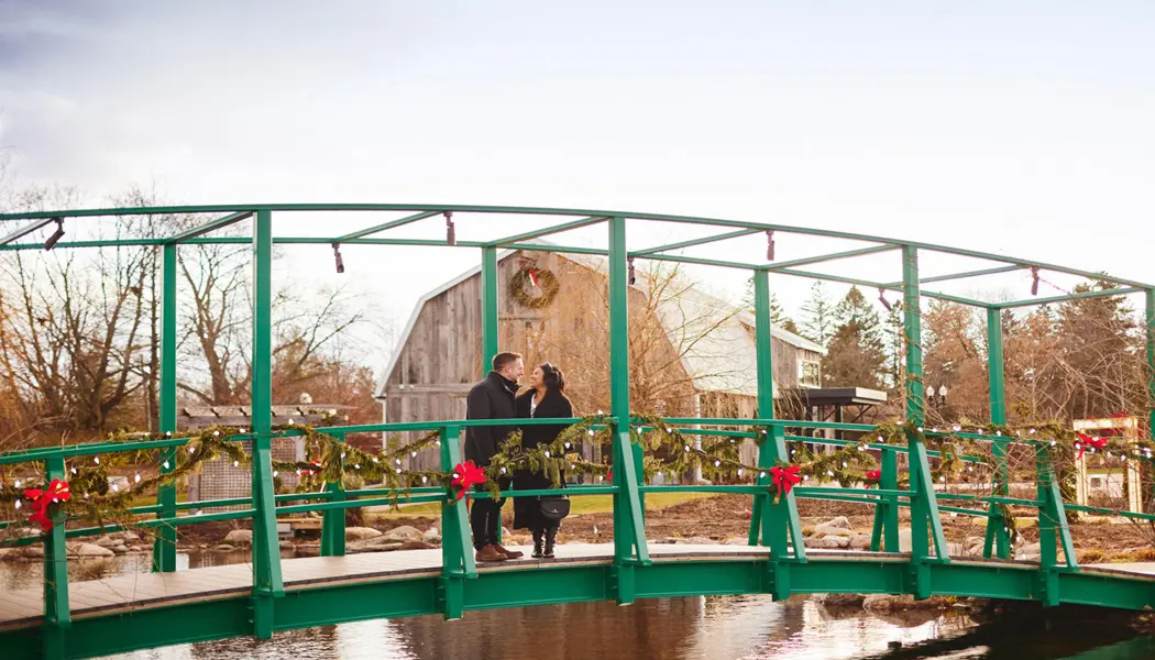 A couple stands on a curved footbridge over a stream decorated with Christmas pine boughs, garlands, bows and lights.