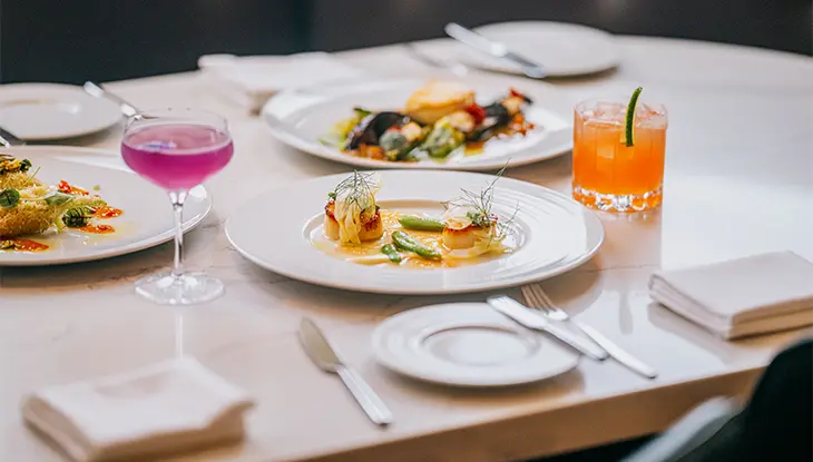 Ein Tisch ist mit drei Gerichten und zwei bunten Getränken gedeckt. Im Vordergrund steht ein Teller mit Jakobsmuscheln und Gemüse in einer reichhaltigen Sauce.